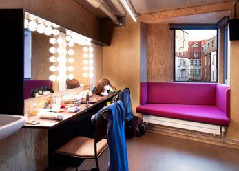2 dressing room table with light up mirrors and a pink sofa underneath a window.