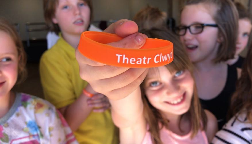 A group of children stand together. In the centre a girl holds up an orange wristband which has the text ‘Theatr Clwyd’ on it.