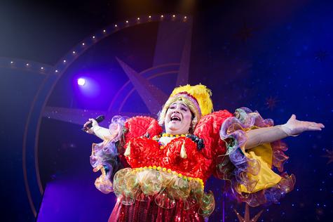 Phylip Harries as the Dame sings with his arms outreached. He is wearing a bright red dress