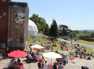 People have fun outside in the sunshine on the grounds of Theatr Clwyd. People from all age ranges are there. Activities include picnics, live music, and haystacks and blowing bubbles for children