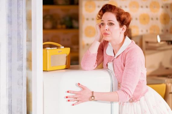 Judy (Katherine Parkinson), dressed in 1950s clothing, leans on her fridge looking dejected. Also on top of the fridge is a yellow radio with a strap