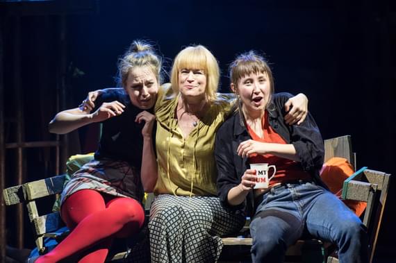A middle-aged woman sits on a bench, her arms around two young women on either side of her. The young woman on the right appears to be mid-speech and is holding a mug saying ‘I love my mum’. The young woman on the left is crying