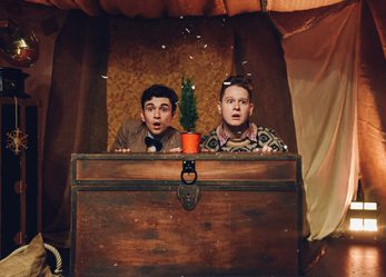 Two young men kneel behind a large chest, looking out over it into the distance in shock. The boy on the left is wearing a tweed jacket with a bowtie while the boy on the left is wearing a patterned jumper. On top of the chest, between the boys, is a small fern tree in a pot