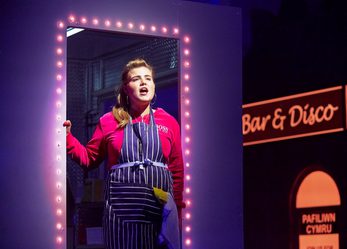 A woman wearing an apron stands in a neon-lit doorway and sings. Behind her a bar/disco is visible