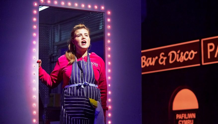 A woman wearing an apron stands in a neon-lit doorway and sings. Behind her a bar/disco is visible
