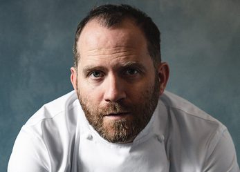 Bryn Williams in Chef uniform, sat posed leaning forwards with his elbows on his knees in front of grey backdrop
