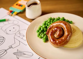 Sausage, mashed potato and peas on a plate on a wooden table top. Colouring paper, a small packets of colouring pencils and a small pot of gravy on the table around it.