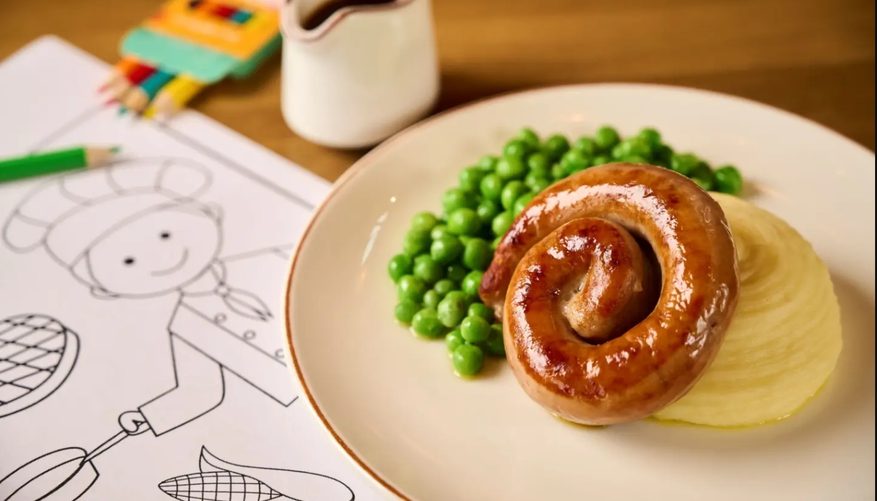 Sausage, mashed potato and peas on a plate on a wooden table top. Colouring paper, a small packets of colouring pencils and a small pot of gravy on the table around it.