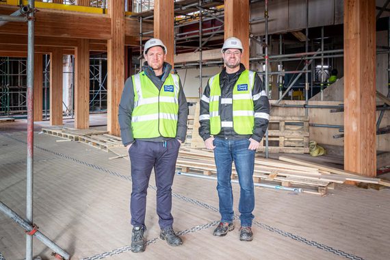 Photo of two men in high vis and hard hats in construction site. They are both part of the senior leadership team, stood in what will be the foyer of our new building.