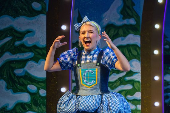 Cinderella stands on stage in our 2025 Panto. She is wearing a blue denim dress with a matching bow in her hair and has her hands raised to her head with an excited expression on her face.