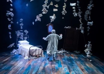 Older man in nightgown facing away from camera on stage with a single bed and grandfather clock. Hundreds of letters dropping from the ceiling.