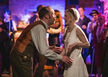 A man and woman at a party, facing each other and smiling in 1920s costumes. In the background there are more partygoers in 1920s costumes.