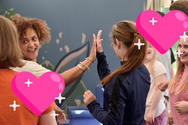 Diverse group of women gathered, two of the women high five in centre of image. 1 large pink emoji heart in the bottom left corner and another on the top right