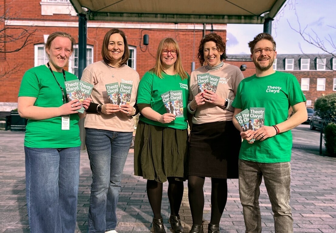 5 Theatr Clwyd company members in light pink or green Theatr Clwyd t-shirts, standing outside a building, holding Theatr Clwyd brochures.