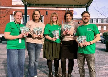 5 Theatr Clwyd company members in light pink or green Theatr Clwyd t-shirts, standing outside a building, holding Theatr Clwyd brochures.