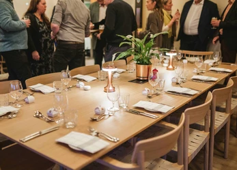 Dining table set with napkins, cutlery, wine glasses, a plant, and table lights. There are people stood around talk behind the table.