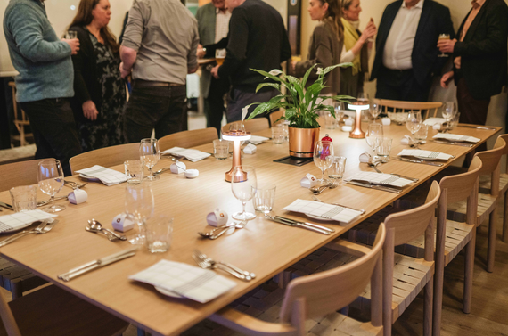 Dining table set with napkins, cutlery, wine glasses, a plant, and table lights. There are people stood around talk behind the table.